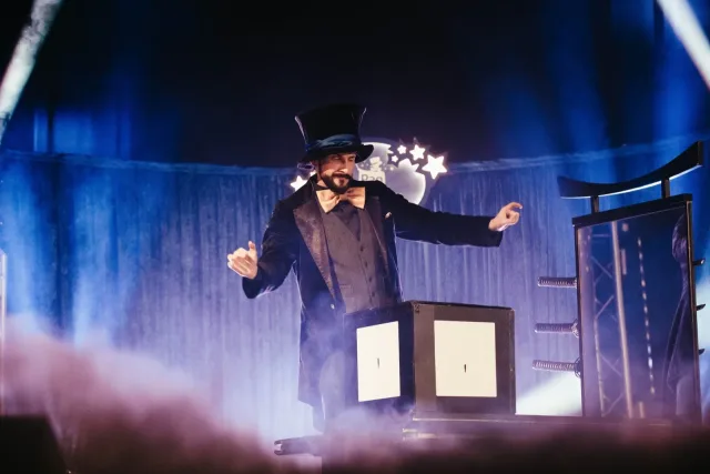 Pan Magiczny w cylindrze przy skrzyni iluzjonistycznej, w dymie i niebieskim świetle scenicznym