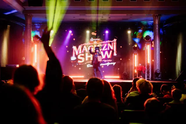 Widownia z uniesionymi rękami przed sceną z neonem Magiczni SHOW